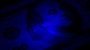 Close-up macro photography of $100 bill security thread glowing under ultraviolet light with metallic details visible, professional currency authentication lighting, dark background