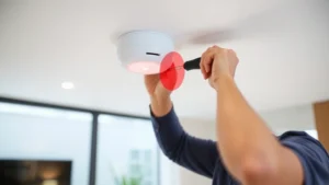 Professional technician installing ceiling-mounted passive infrared motion sensor in modern home interior, using mounting bracket and screwdriver, showing sensor lens and detection area